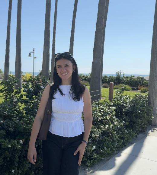 Portrait photo for Kayla Joachin in a white top and dark pants with palm trees in the background