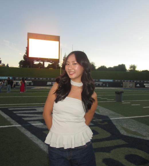 Portrait photo of Laura Zhou in a white top and blue jeans with a football stadium in the background