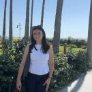 Portrait photo for Kayla Joachin in a white top and dark pants with palm trees in the background