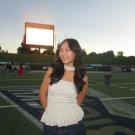 Portrait photo of Laura Zhou in a white top and blue jeans with a football stadium in the background